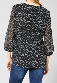 Blouse noire à pois gris, manches semi-transparentes, décolleté arrondi et poignets élastiques. Associée à un jean bleu.