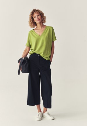 Kobieta w limonkowym topie z dekoltem w kształcie litery V, czarne szerokie spodnie do połowy łydki, białe trampki, trzyma czarną torbę, stoi na jednolitym tle.