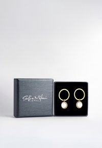 Guldörhängen i form av hoops med en cirkulär genomskinlig sten, presenterade i en svart texturerad smyckeslåda med vit branding.