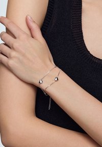 Srebrna bransoletka z dwoma sercowymi zawieszkami. Posiada delikatny łańcuszek i regulowaną długość, noszona na nadgarstku w zestawieniu z ciemnym swetrem.