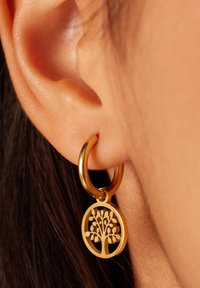 Gouden oorbellen in de vorm van een ring met een uitgesneden boomontwerp als hanger. De ring is glad en glanzend, terwijl de hanger ingewikkelde takdetails heeft.