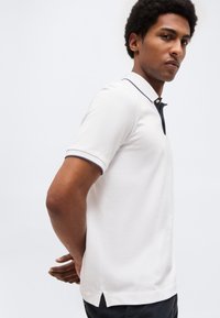 Νέος άνδρας με σταυρωμένα χέρια πίσω από την πλάτη, φορώντας λευκό πόλο μπλουζάκι με κοντά μανίκια και σκούρο περίγραμμα στο γιακά και τα μανίκια.