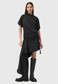 Top nero fluido con scollo drappeggiato, abbinato a una gonna asimmetrica. La modella indossa stivali neri fino al ginocchio con dettaglio in zip dorato.