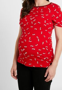 Röd kortärmad graviditetstopp med små vita blommönster. Mjukt, elastiskt tyg med rund hals och svart kant.