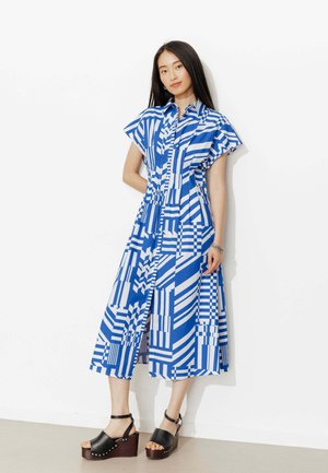 Femme portant une robe midi à motifs géométriques bleus et blancs avec des manches courtes et des sandales à plateforme noires, debout contre un mur blanc.