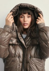 Brązowa kurtka puchowa o błyszczącej fakturze, z dużym kapturem, zatrzaskami oraz bocznymi kieszeniami. Wyściółka zapewnia ciepło i komfort.