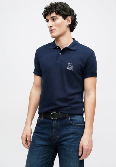 Μπλε σκούρο πόλο μπλουζάκι με κοντά μανίκια διπλωμένα, με λογότυπο EA στο στήθος. Φορεμένο με σκούρο μπλε τζιν και μαύρη ζώνη.