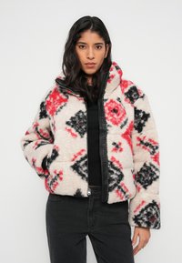 Φλις μπουφάν σε μπεζ χρώμα με μαύρα και κόκκινα γεωμετρικά σχέδια, εμπρός με φερμουάρ, ψηλό γιακά και ελαστικές μανσέτες. Μαλακή υφή.