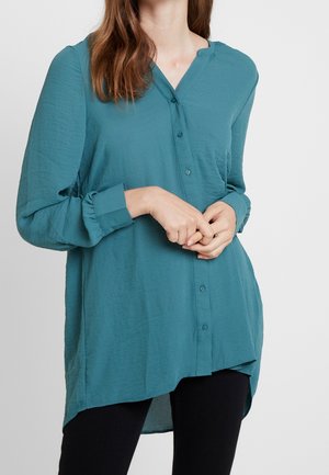 Camicia a maniche lunghe color teal con chiusura a bottoni, vestibilità comoda e orlo curvo. Realizzata in un tessuto leggero e strutturato.