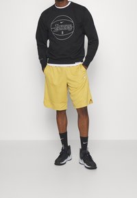 Sudadera negra con logo blanco de "Lakers", combinada con pantalones cortos deportivos amarillos. Zapatillas y calcetines negros completan el conjunto.