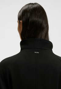 Frau mit langen dunklen Haaren, die eine schwarze Fleecejacke mit hohem Kragen und kleinem silbernen "BOSS"-Logo am Nacken trägt.