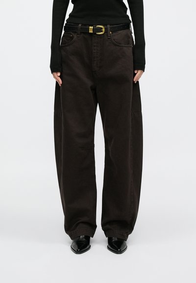 Calças castanhas de perna larga feitas de algodão, com um cinto preto de fivela dourada e detalhes subtis de costura. Usadas com sapatos pretos.