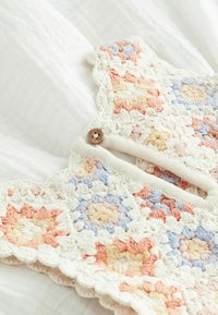 Robe pour bébé au crochet avec des carrés granny en couleurs pastel, des bords en feston et un bouton en bois sur un fond de tissu texturé blanc.