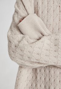 Lys beige strikket sweater med et struktureret mønster, der har ribbede manchetter og en oversize pasform, som viser åbne vævedetaljer gennem hele designet.