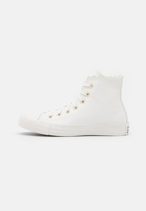 Witte hoge canvas sneaker met een omboorde rand, gouden veterogen, witte veters en een rubberen zool op een effen witte achtergrond.
