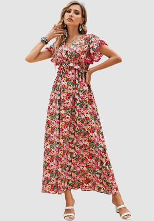 Mujer que lleva un vestido largo floral colorido con mangas con vuelo y ribete rosa, complementado con pulseras, pendientes y sandalias blancas de tacón.
