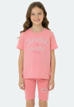 Fille souriante aux longs cheveux ondulés portant un ensemble t-shirt et short rose assortis Lee Cooper avec le texte East London.