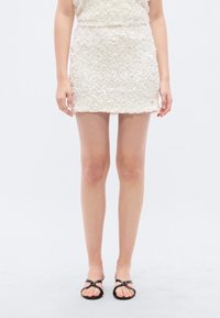 Λευκή, υφασμάτινη μίνι φούστα με στρώσεις διακοσμήσεων που μοιάζουν με πέταλα. Φορεμένη με μαύρα σανδάλια πλατφόρμας με λουριά και μικρή πόρπη.