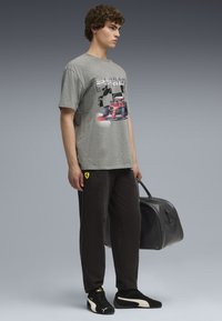 Ung mand iført grå Scuderia Ferrari T-shirt, sorte bukser med Ferrari-logo, sorte Puma-sneakers, mens han holder en sort læder sportstaske.