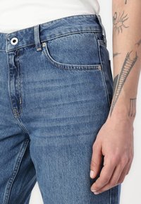 Mėlynos džinsinės kelnės turi standartinį pritaikymą su išblukusiu tekstūriniu paviršiumi, metalinę sagą ir priekinėmis kišenėmis. Tatuiruota ranka laiko juosmens diržą.