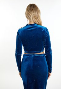 Costum din catifea albastră format din două piese, cu un top scurt cu mâneci lungi și nasturi pe umăr și o fustă până la podea cu o textură netedă.
