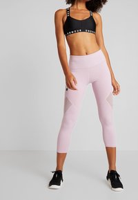 Svart sportbh med logoband, ihop med ljusrosa capribyxor med meshpaneler och en åtsittande design. Svarta träningsskor.