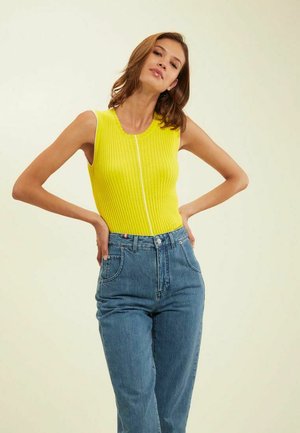Haut à côtes sans manches en jaune vif avec une fermeture éclair à l'avant. Associé à un jean en denim bleu taille haute. Texture lisse, coupe décontractée.