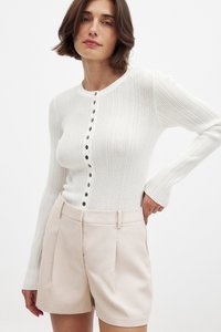 weißes, geripptes Langarmoberteil mit schwarzen Knöpfen in der Mitte, kombiniert mit hellrosa maßgeschneiderten Shorts. Glatte Texturen, figurbetontes Design.