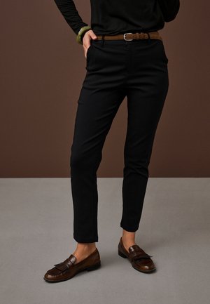 Person iført sorte slim-fit bukser, brun læderbælte, mørkebrune loafers, sort langærmet top og grøn armbånd med hånd i lommen.