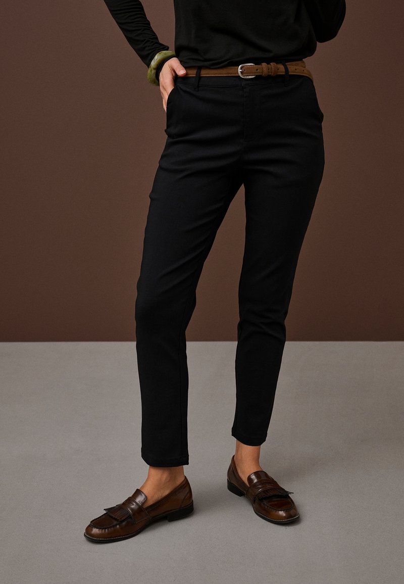 Personne portant un pantalon noir slim, une ceinture en cuir marron, des mocassins marron foncé, un haut noir à manches longues, et un bracelet vert, la main dans la poche.
