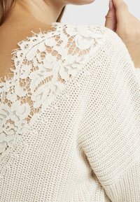 Nahaufnahme einer Person, die einen cremefarbenen Strickpullover über einem weißen Spitzenoberteil mit Blumenmuster trägt, wobei Schulter- und Handdetails sichtbar sind.