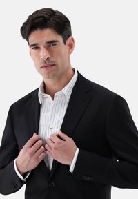 Schwarze Anzugjacke über einem weißen Hemd mit senkrechten Streifen. Die Jacke hat einen Reverskragen und einen einfachen Knopf. Hände greifen die Revers der Jacke.