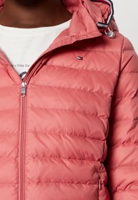 Różowa puchowa kurtka z zapięciem na zamek, wyposażona w poziome przeszycia oraz kaptur. Zawiera małe logo na lewej piersi oraz akcenty z gumką.