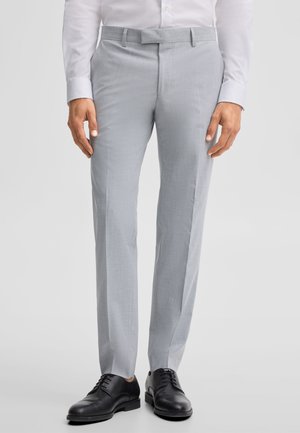 Man die lichtgrijze nette broek, wit overhemd met lange mouwen en zwarte nette schoenen draagt, staande op een lichtgrijze achtergrond.