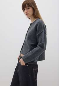 Cardigan in lana grigia con scollatura rotonda, chiusura frontale con bottoni e vestibilità rilassata. Indossato sopra jeans neri a vita alta con tasche.
