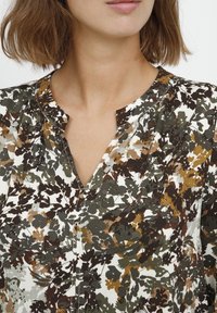 Blusa floreale bianca con motivi verdi, marroni e gialli. Presenta una scollatura a V e un tessuto leggermente testurizzato. Maniche corte, vestibilità comoda.