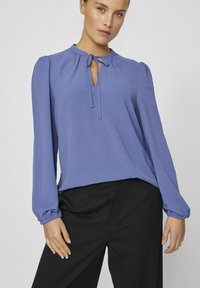 Blusa azzurra chiara con scollo a chiave, maniche lunghe a sbuffo e polsini elastici. Indossata con pantaloni neri su misura. Tessuto morbido.