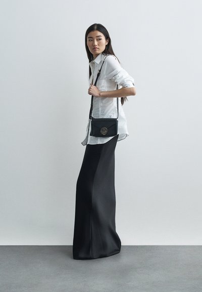 Femeie îmbrăcată cu o bluză albă și o fustă lungă neagră, ținând o geantă crossbody neagră cu un simbol de pace zăpadă, stând pe un fundal simplu.