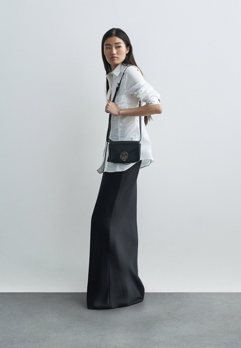 Nő fehér blúzban és hosszú fekete szoknyában, fekete keresztpántos táskával, amelyen arany békeszimbolum van, síma háttér előtt áll.