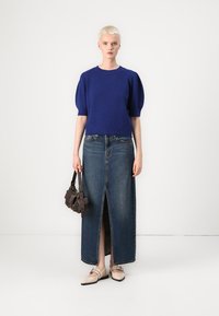 Un pull en tricot bleu à manches bouffantes associé à une longue jupe en jean avec une haute fente, complété par des chaussures plates beiges et un petit sac à main marron.