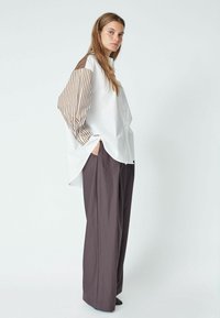 Žena stojí s rukami v vreckách, oblečená v bielej oversized košeli s hnedými prúžkovanými rukávmi a voľných hnedých nohaviciach na jednolatom pozadí.