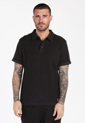 Man met kort haar en tatoeages op beide armen draagt een zwart shirt met korte mouwen en een kraag, en zwarte broek, staand tegen een witte achtergrond.