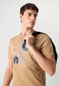 Ung mann med krøllete hår har på seg en beige t-skjorte med grafikk og bærer en svart ryggsekk over den ene skulderen, og ser mot siden mot en ensfarget bakgrunn.