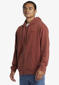 Hnedočervená zipsová mikina s veľkou kapucňou, sťahovacími šnúrkami, klokaními vreckami a malou logom na ľavej strane hrude. Mäkká textúra látky.