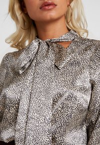 Blusa di seta caratterizzata da una base beige chiaro con motivo a macchie nere, dettaglio a fiocco al colletto e texture morbida e liscia.