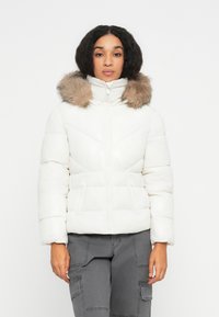 Vrouw in een witte pufferjack met een met bont omzoomde capuchon en grijze cargobroek, staande tegen een effen witte achtergrond.