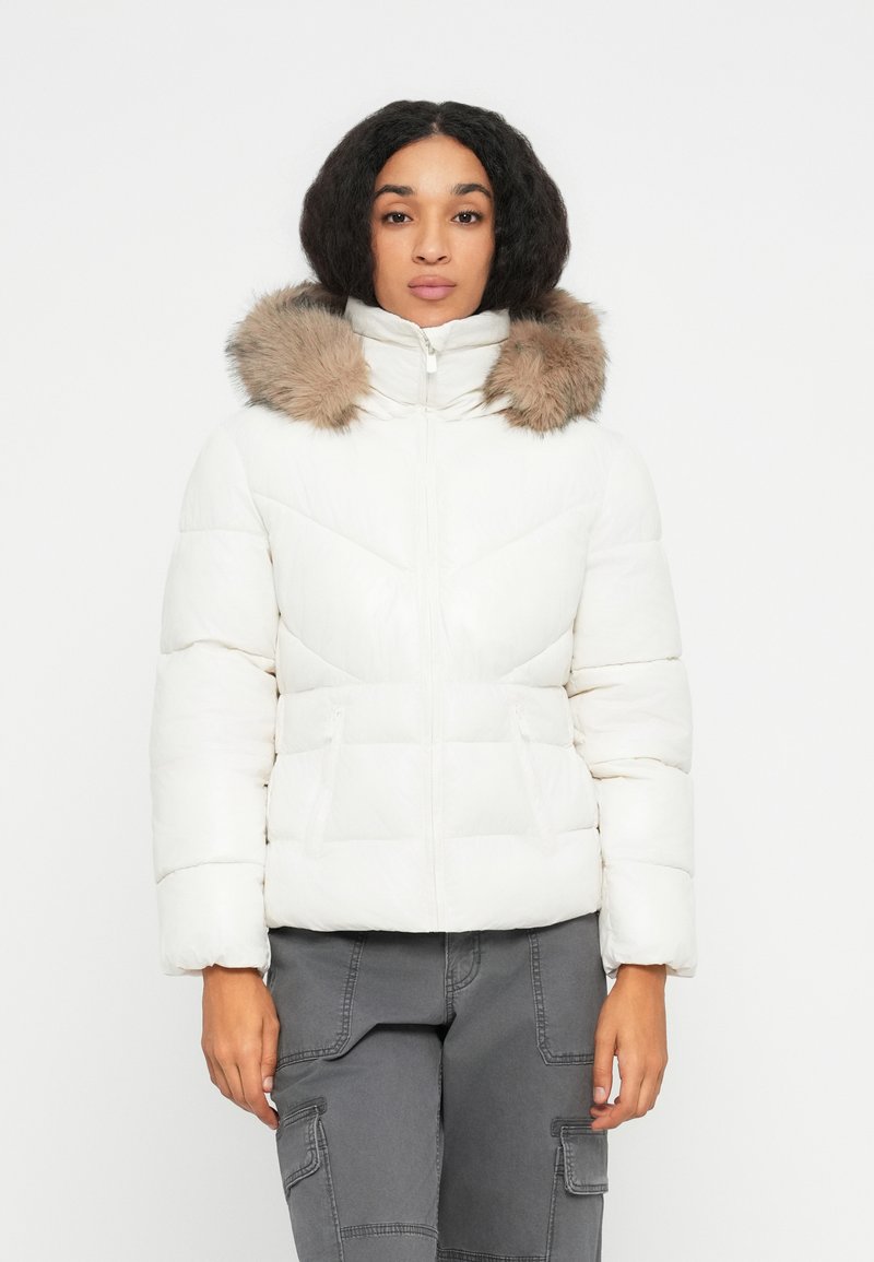 Vrouw in een witte pufferjack met een met bont omzoomde capuchon en grijze cargobroek, staande tegen een effen witte achtergrond.