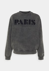Sudadera de forro polar gris con cuello redondo. Presenta un gran texto negro "PARÍS" en la parte frontal y un pequeño parche de logo en la manga izquierda.