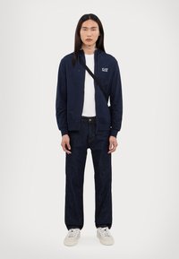 Marineblå zip-up hettegenser med logo, hvit t-skjorte under, mørk denimjeans og beige sneakers, båret av en stående modell mot en ensfarget bakgrunn.