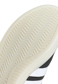 Suola di una scarpa con materiale in gomma bianca, caratterizzata da un motivo a zigzag e spille bianche e nere lungo il lato.